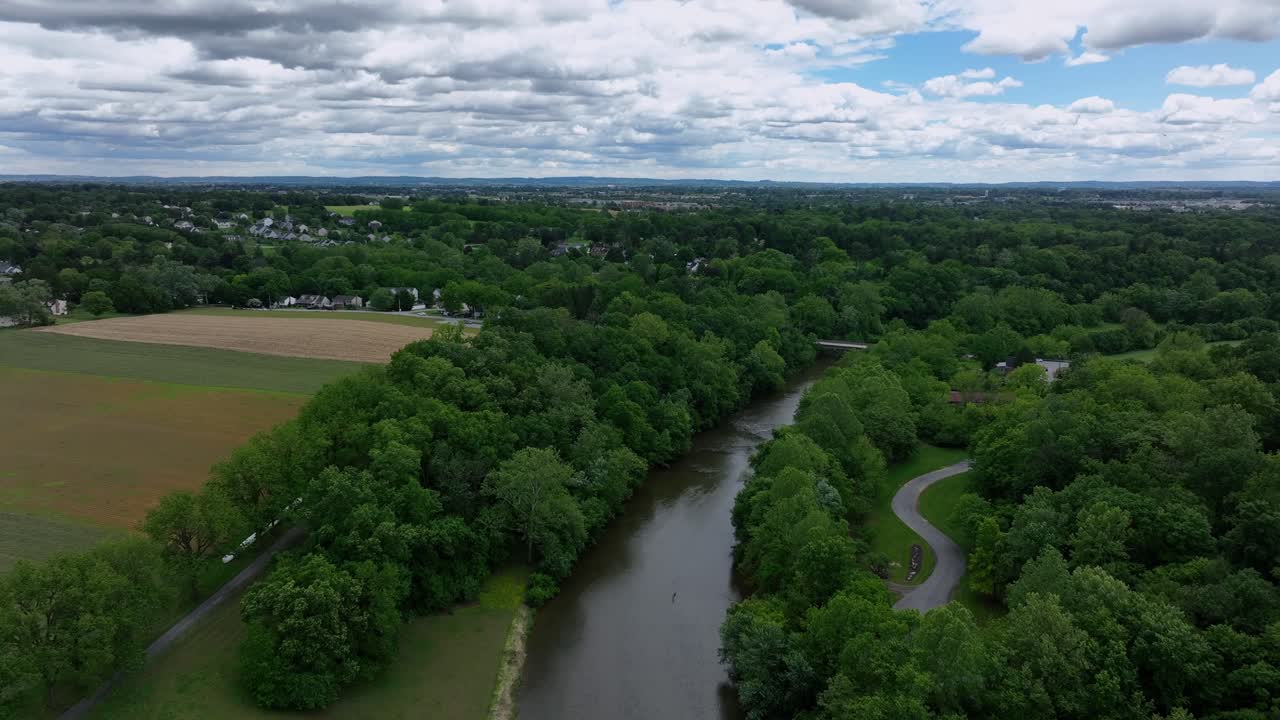 River in rural area of american city. Aerial rising wide shot. Forest trees and path between agricultural landscape. Houses and homes in distance. Cloudy sky in spring season.