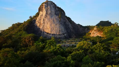 4k-Low-Aerial-Pan-above-treetops-towards-the-Buddha-Mountain-in-Pattaya-at-dawn,-golden-light-on-the-image-of-Buddha-in-Khao-Chi-Chan,-Chonburi,-Thailand