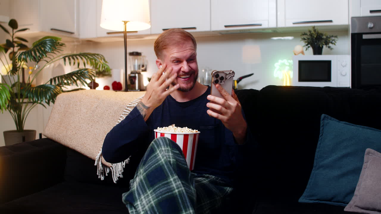 Ung mand slapper af i sofaen med sin smartphone, skifter kanal, griner, spiser popcorn og nyder en stille aften derhjemme.