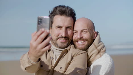 front view of a happy gay couple taking selfie on phone on the beach