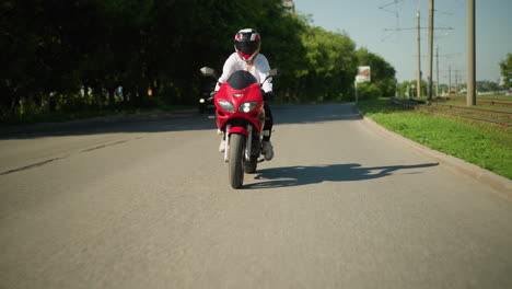 a female motorcyclist wearing a helmet rides fast on a red power bike along a road, the background shows a blur of lush green trees and a car approaching from behind