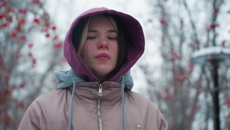 close up of young lady adjusting her hoodie cap while looking directly at the camera with serious expression, standing outdoors in winter jacket, blurred trees background