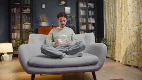 young man sitting on sofa wearing wireless headphones and watching some shocking movie scene on smartphone