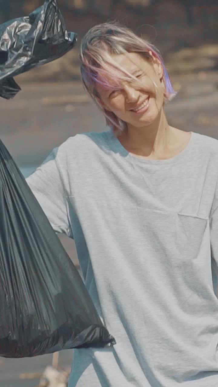 Young caucasian woman eco volunteer with garbage shows thumb up stands on beach