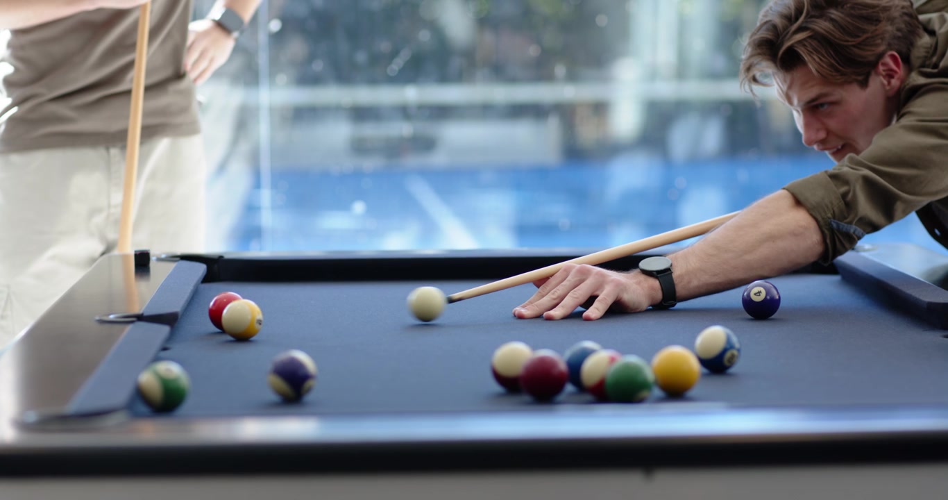Male friends playing pool by glass window, primary player lining shot, striking cue to pocket balls