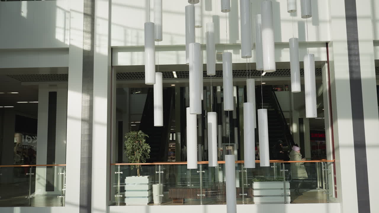 Contemporary indoor view of shopping mall with people walking past elegant hanging lights and glass railings, while escalators lead to upper floor surrounded by sleek white architecture