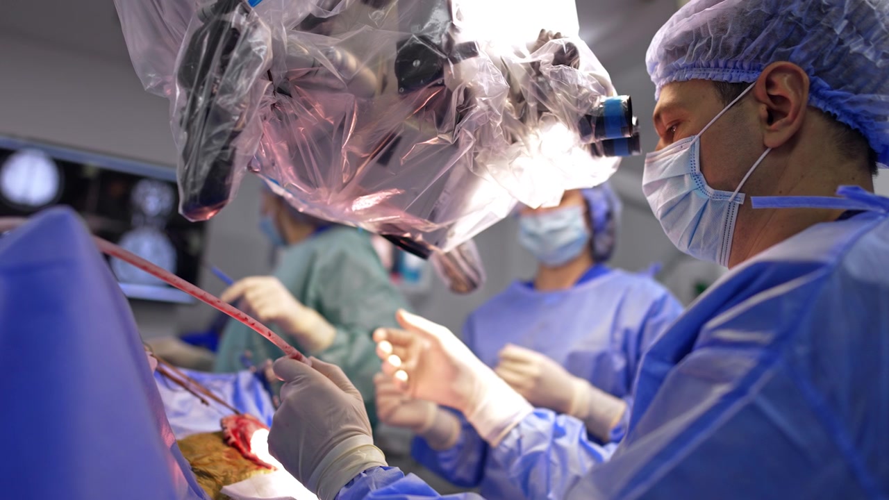 En professionel neurokirurg sidder ved mikroskopet og bruger et sugeinstrument. Assistenter giver instrumentet til lægen.