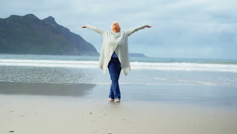 Senior-woman-standing-with-hands-raised-on-beach