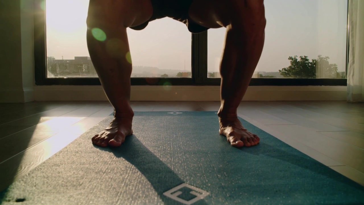 Un atleta si piega abbassando i fianchi su un tappetino blu vicino a una finestra alta mentre la luce del sole si irradia, indossando pantaloncini scuri e a piedi nudi.