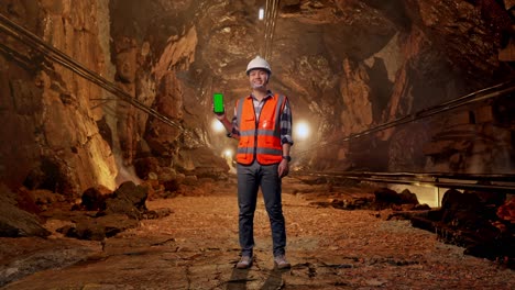 full body of asian male engineer with safety helmet smiling and showing green screen smartphone to the camera while standing in underground mine tunnel