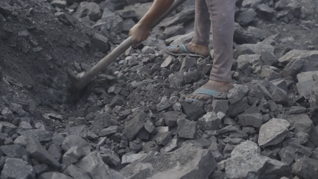 Young male worker extracting coal at coal mine, wearing open toed sandal and pant, the individual is actively engaged in breaking or moving jagged pieces of coal using a hand tool, Bokaro, Jharkhand