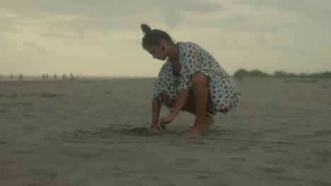 An-Indian-woman-kneels-in-the-soft-sand,-her-hands-delicately-tracing-patterns-on-the-beach