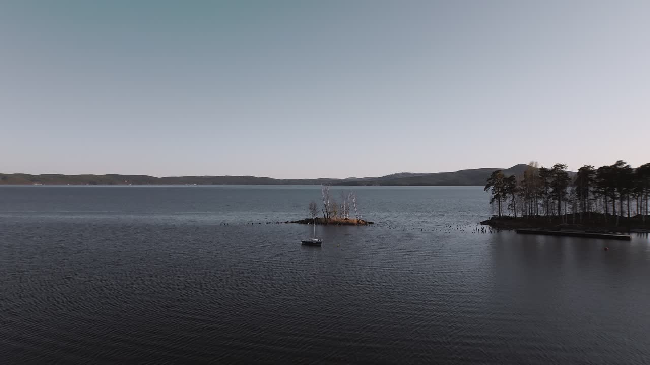 Drohnenaufnahme des ruhigen Turgoyak-Sees mit einer kleinen bewaldeten Insel und einem einsamen Segelboot auf dunklem Wasser in der Dämmerung, Landschaft des Uralgebirges, Oblast Tscheljabinsk, Russland