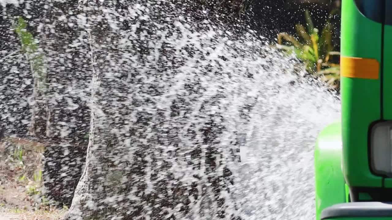 A green truck sprays water onto roadside trees, creating a refreshing splash effect.