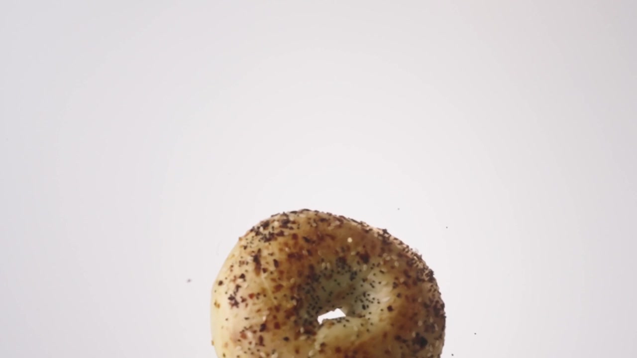 Everything-seeded savory bagels raining down on white seamless backdrop in cinematic slow motion. new york deli branding, breakfast sandwich ads, bagel shop content.