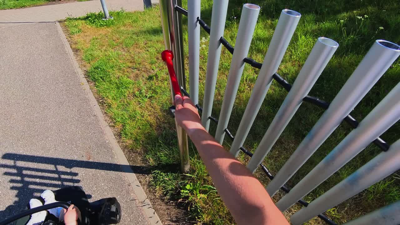 A person playing outdoor tubular bells with a red mallet, set in a grassy park environment. Ideal for themes related to music, outdoor activities, interactive play, and sound exploration.