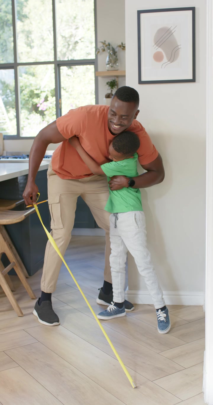 Vertical video: African American dad entering home, measuring floor with tape, high-fiving, hugging