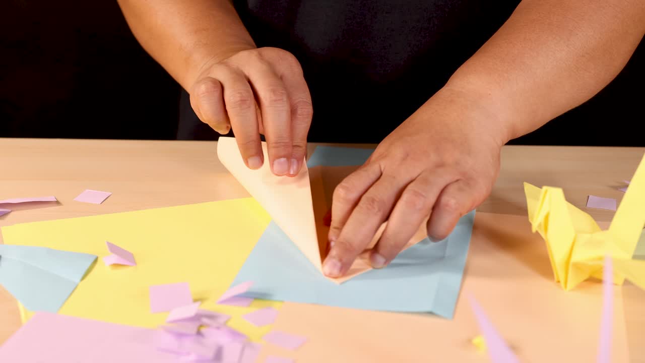 Person folds pastel origami paper crane on wooden table under warm, even studio lighting