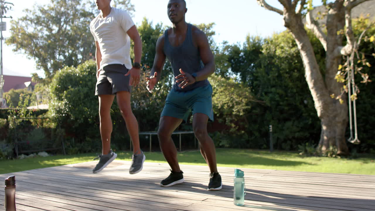 Exercising outdoors, two multiracial male friends jumping on wooden deck with water bottle nearby