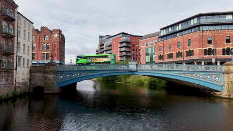 a green double-decker bus travels across a blue victorian bridge spanning the river aire, framed by historic red-brick buildings under overcast daylight. static wide shot