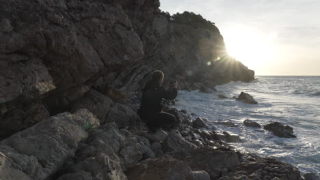 hombre en cuclillas tomando una foto del sol de la mañana sobre el mar, se aleja a lo largo del acantilado