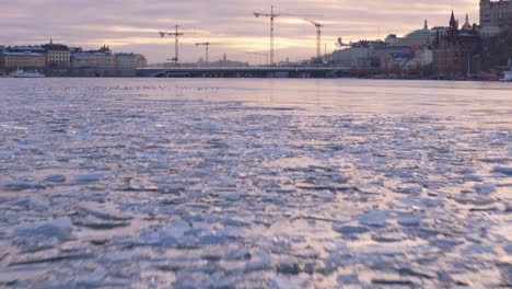 Low-aerial-tilt-up-reveal-over-frozen-Riddarfjärden-at-sunset-toward-Slussbron