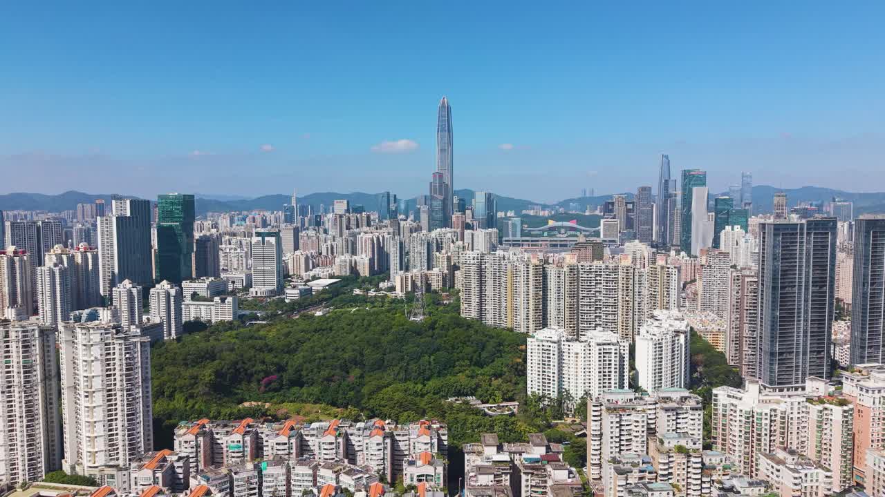 Aerial view of Shenzhen’s skyline featuring Ping An Finance Centre rising above dense urban districts. Modern high-rises, green park areas, and distant mountains under a clear blue sky. China, UHD