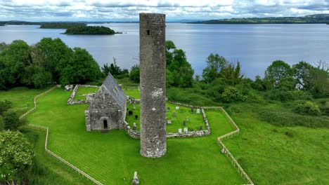 Statische-Drohnenaufnahme-Von-Rundem-Turm-Und-Kirche-Mit-Lough-Derg-Im-Hintergrund,-Heilige-Insel,-Wichtige-Historische-Religiöse-Stätte-Am-Shannon-Fluss,-Irland,-Epische-Orte