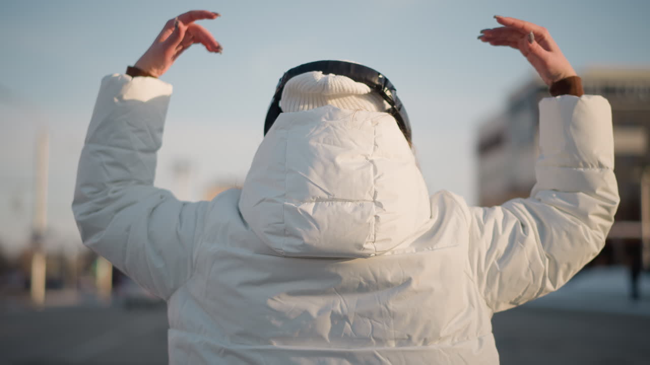 Cheerful dancer wearing white winter coat and beanie moves rhythmically with raised arms in open urban space, back turned to camera, enjoying winter atmosphere under clear sky