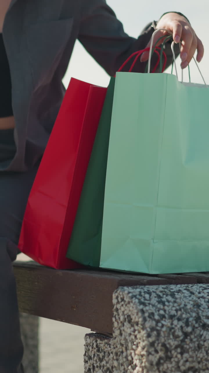 dama en ropa gris cae verde, menta, bolsas de compras rojas en el banco mientras está sentado, con una ligera vista de la colina de vegetación en el fondo, y el objeto borroso de distancia