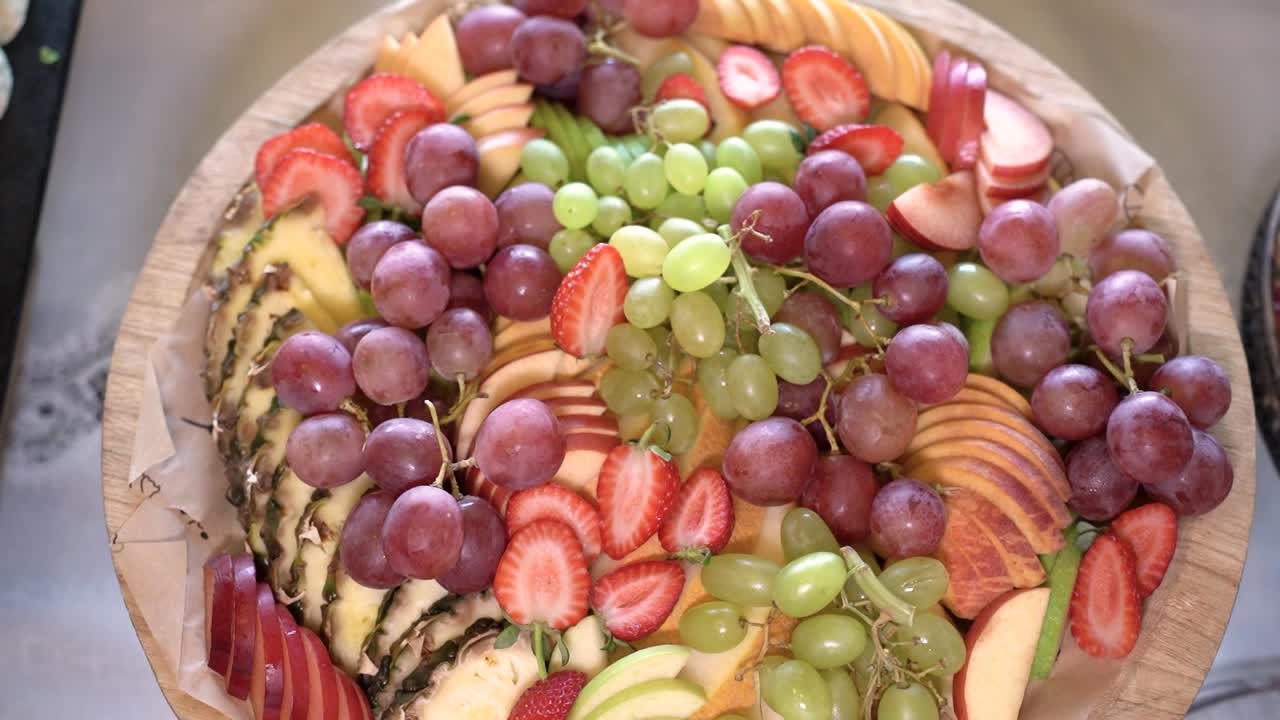Et stort, rundt fat er fylt med et utvalg av frisk frukt. Ulike typer druer, jordbær, ananas og epler er pent arrangert.