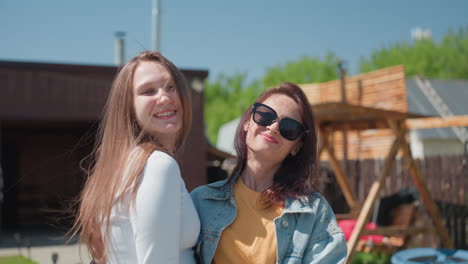 two happy women embrace warmly in sunlit backyard, smiling brightly in front of wooden house and garden, blurred background shows another girl lying on swing