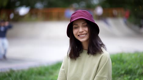 Portrait-of-a-brunette-girl-in-a-green-sweater-in-a-hat-sits-against-the-backdrop-of-a-skatepark-and-poses-for-the-camera.-Walk