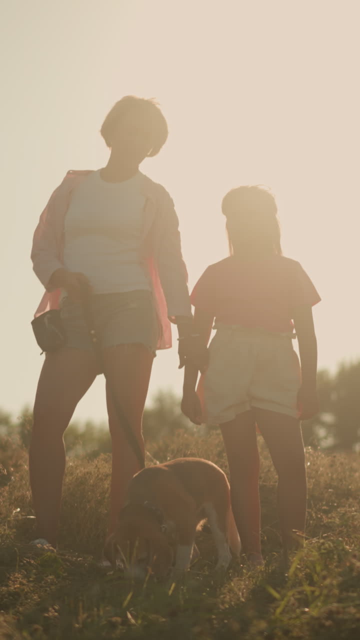 Family standing on hilly farmland during sunset with sun shining behind them as mother reach out for her daughter hand, featuring two women and child with dog