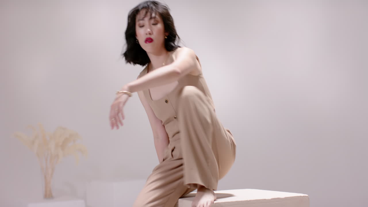 After pause Asian woman entering studio by block sliding onto beige cube showing beige outfit