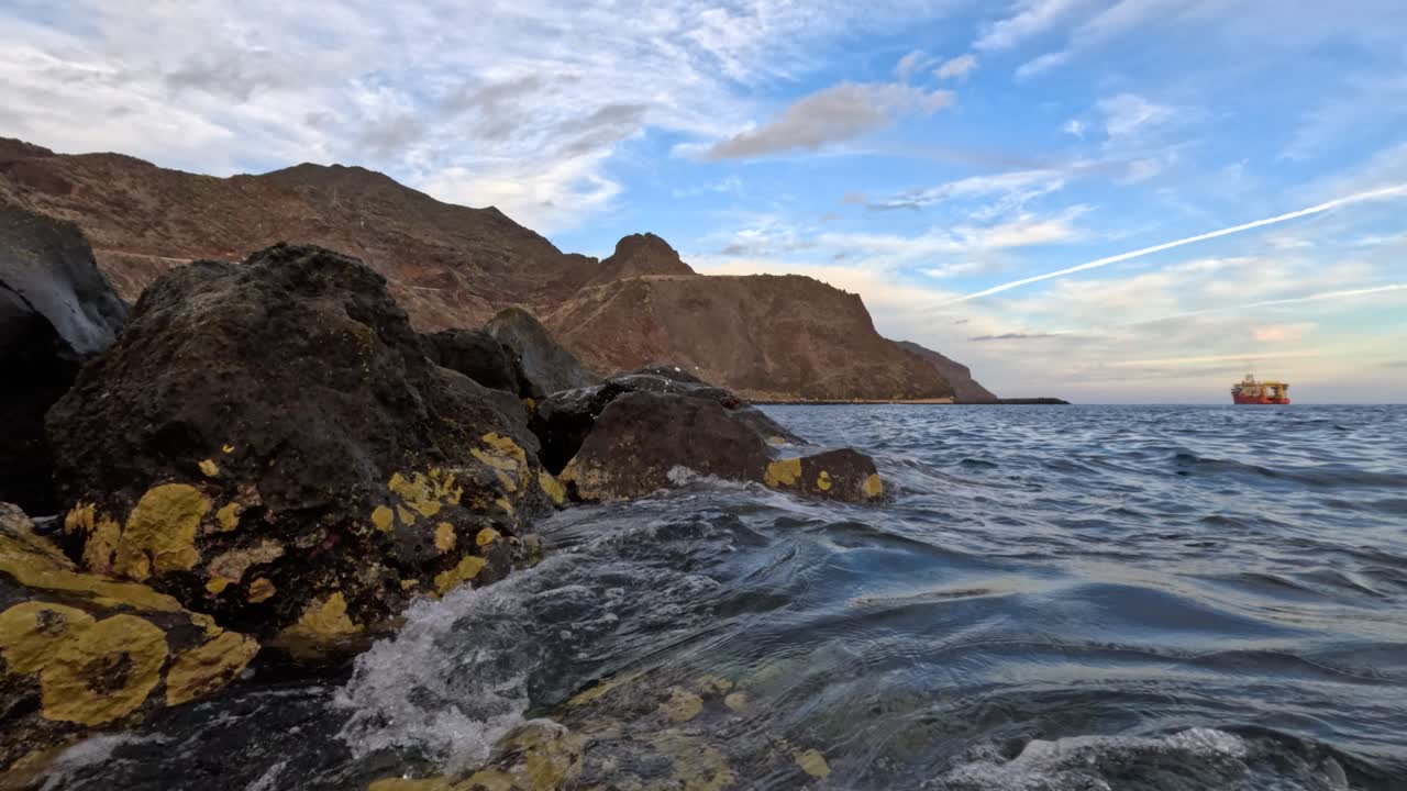 fotografía de cerca de las rocas con musgo y olas salpicando contra ellas en la costa de tenerife con un barco de carga en el fondo