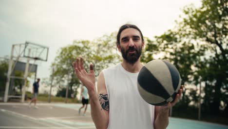 a middle-aged man in a white t-shirt with a beard poses against the background of a basketball court with a basketball in his hands