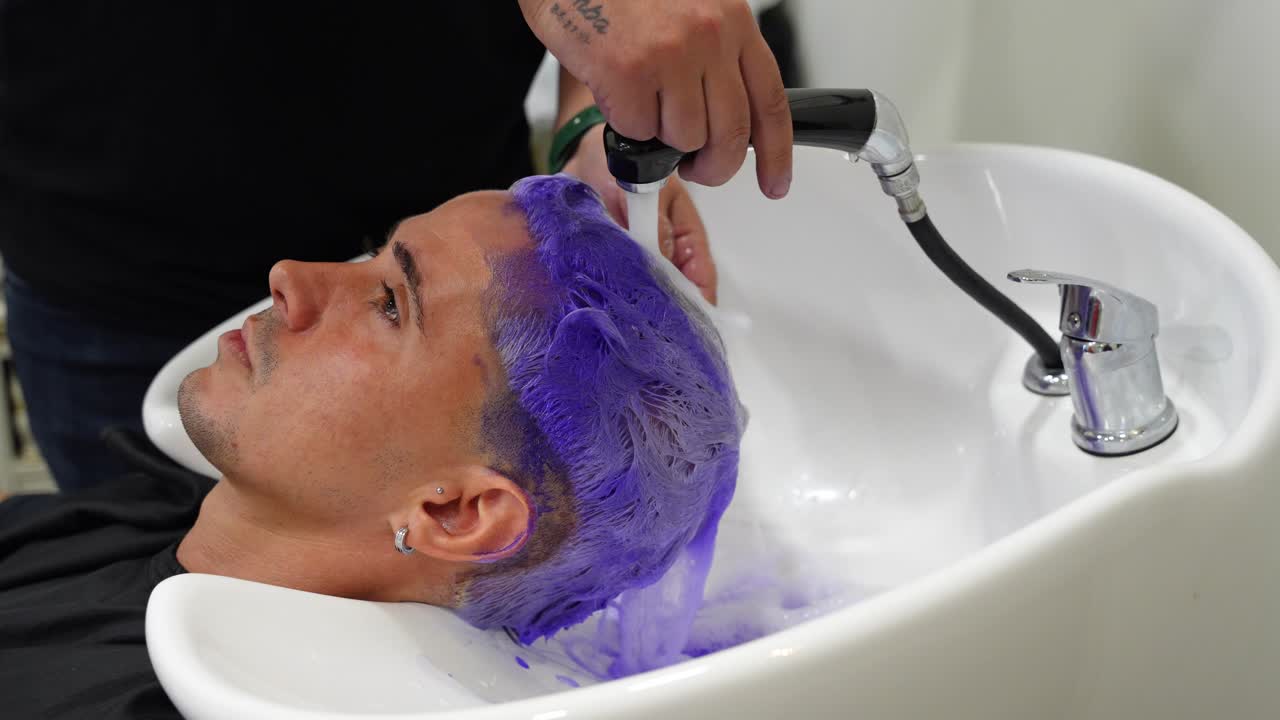 Primo piano di un parrucchiere che risciacqua lo shampoo viola dai capelli di una cliente sotto l'acqua del lavandino durante il processo di decolorazione