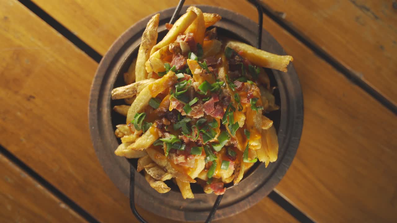 A stationary top-down wide-angle shot of a full bowl of bacon and french fries coated with cheddar cheese and topped with spring onions
