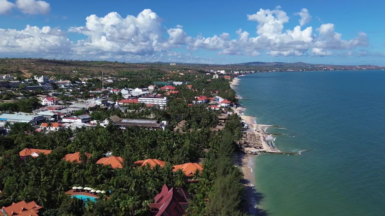 Cảnh quay flycam ghi lại nét đặc trưng nhiệt đới của Mũi Né, Việt Nam, với thảm thực vật dừa dày đặc, các tòa nhà dân cư, những mái nhà đầy màu sắc và làn nước biển xanh biếc trải dài dọc bãi biển