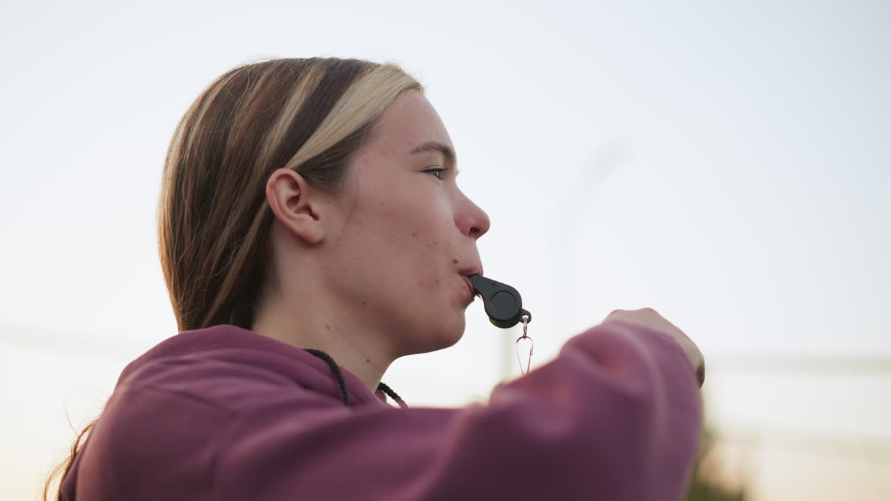 뒷면에 나무와 환경이 흐릿한 상태에서 사람들이 훈련하는 동안 입에 휘파람을 휘두르는 강사의 클로즈업으로 야외 배구 코칭을 보여줍니다.
