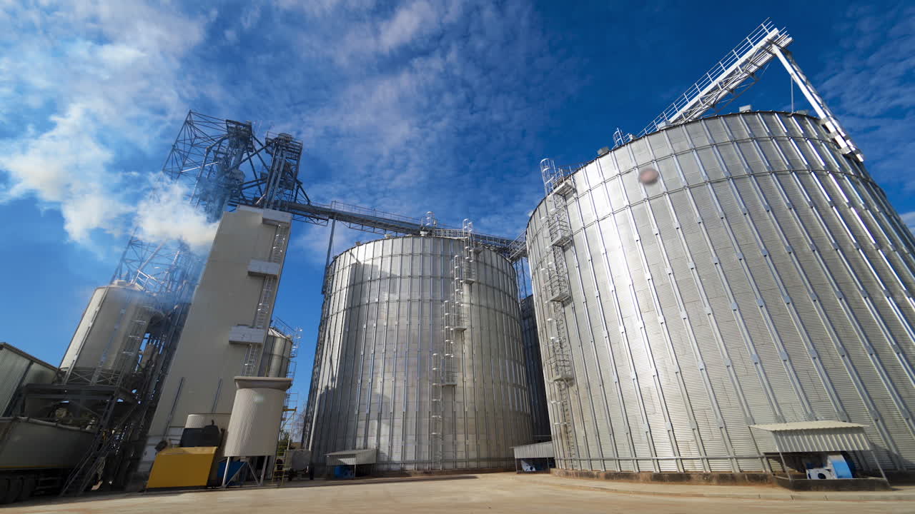 Harmful smoke filling the atmosphere from industry. Aluminum grain elevators on the modern agricultural factory at bright sunny day.