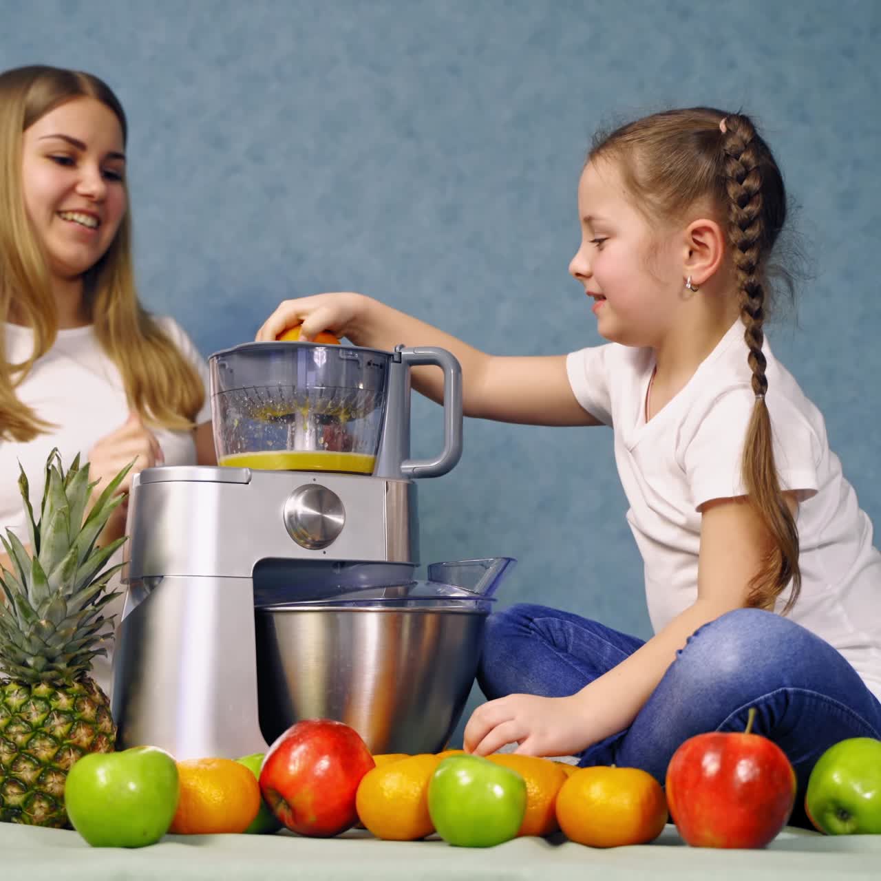 Little girl and mother near the squeezer. Fresh organic fruits on the table. Cute small girl eating orange while sitting on the table full of ripe fruits.