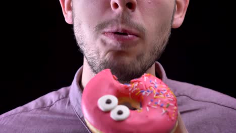 close-up portrait of man biting and chewing delicious glazed pink donut with multicolored chips and eyes on black background.