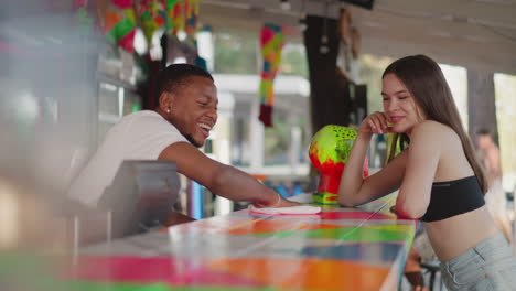 barman flirts with young woman at counter in beach bar. black waiter wipes colorful countertop smiling to pretty lady in club. bartender entertains guest