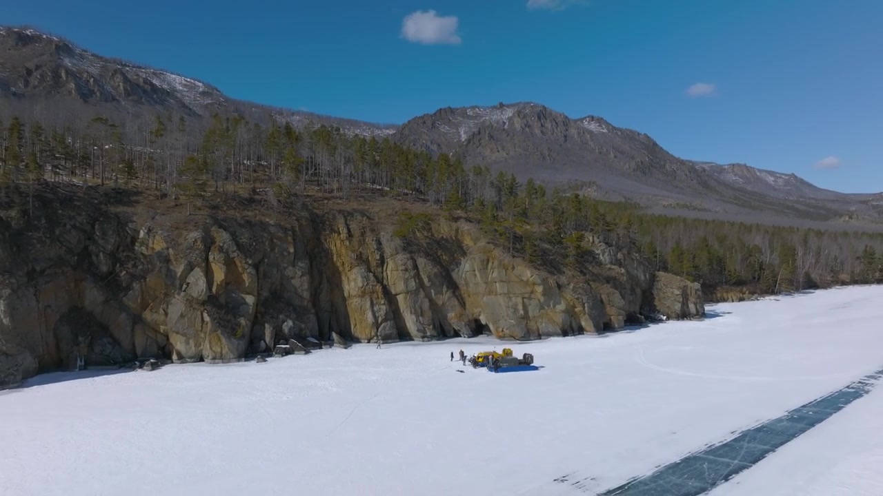 Riprese drone del Lago Baikal ghiacciato con costa rocciosa, foresta di taiga e cresta montuosa sotto un cielo blu, paesaggio invernale nell'Oblast di Irkutsk, Russia.