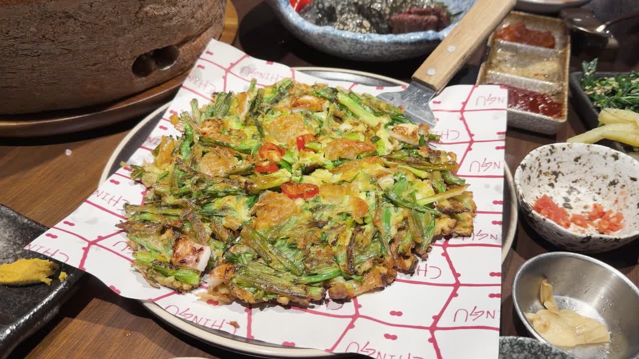 Hand slices and serves Korean vegetable omelette on wooden table with side dishes, overhead view