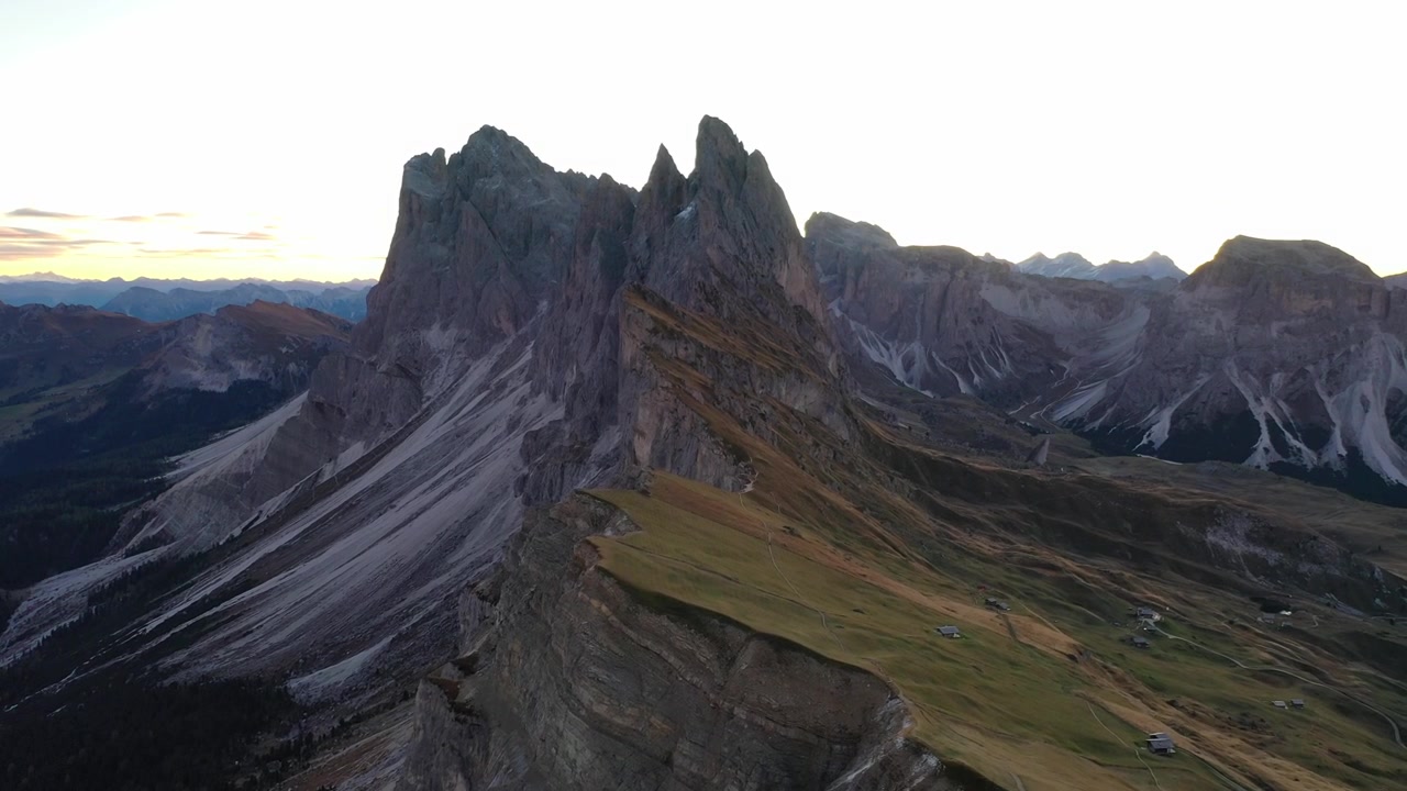 Breathtaking high-angle drone shot of the dramatic Seceda peak and sloped alpine meadows in Val Gardena, Italy. Features majestic jagged mountain ridges illuminated by warm golden hour sunset light.