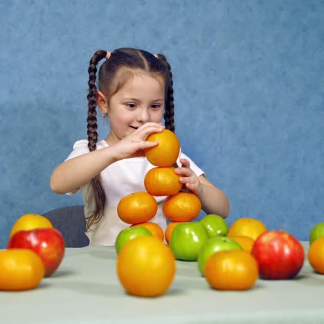 cute little girl with fresh fruit. funny girl building a tower of oranges on the table. child playing with organic fruit on blue background.