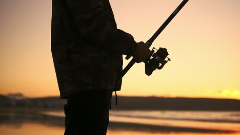 man silhouette handle rotation with reel of fishing rod against of orange sunset slow motion